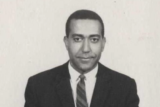 A black and white photo of a person with short dark hair, wearing a black suit and dark tie